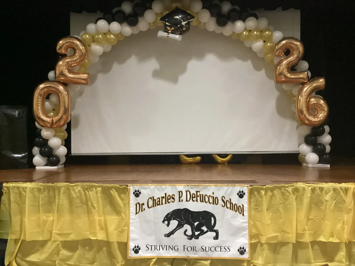 Black, white and gold balloon arch with gold 2026 foil numbers framing a school stage