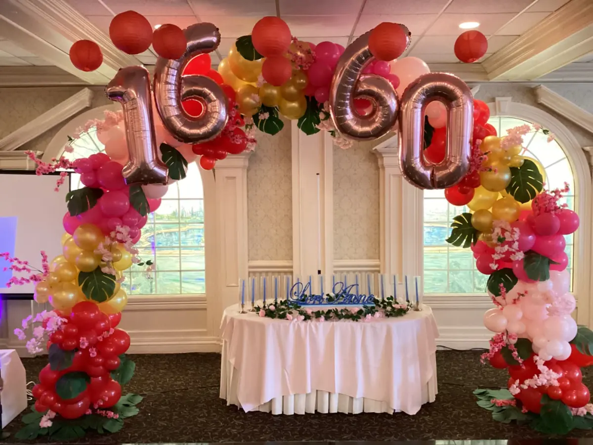 Organic pink, red and gold balloon arch with tropical leaves and rose gold 16 and 60 foil numbers over a head table, New Jersey