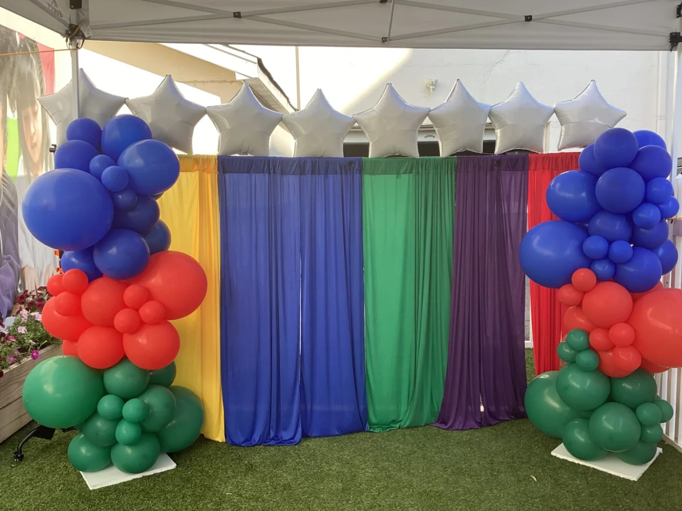 Organic blue, red and green balloon columns with silver star foils framing a rainbow drape backdrop