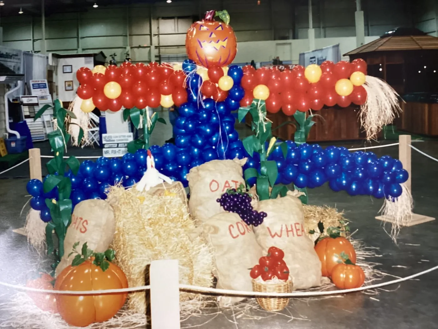 Balloon scarecrow in red, yellow and blue with a jack-o-lantern head, surrounded by hay bales, grain sacks and orange balloon pumpkins at a harvest display, New Jersey